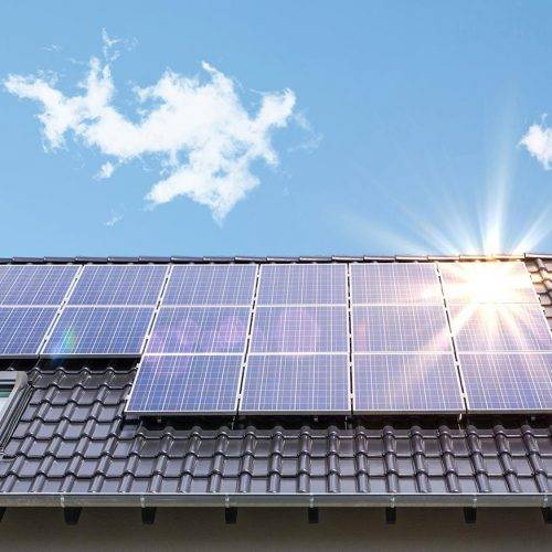 Die Dachfläche eines Wohnhauses, auf der mehrere Solarmodule installiert sind. Die Solarpaneele sind gleichmäßig angeordnet und fangen direktes Sonnenlicht ein, wobei auf der rechten Seite ein heller Lichtreflex die Energie der Sonne symbolisiert. Der Himmel ist klar mit wenigen kleinen Wolken, was auf gute Wetterbedingungen und optimale Solareinstrahlung hinweist. Die Dacheindeckung besteht aus dunklen Dachziegeln, und unter den Modulen ist ein Fenster in das Dach integriert.