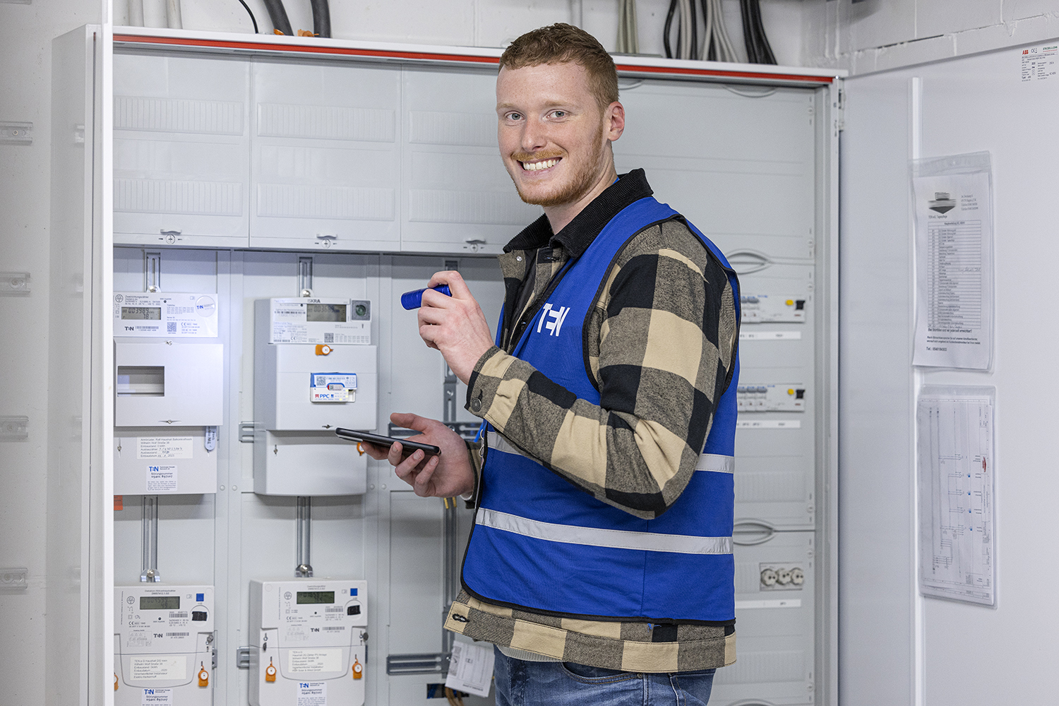 Junger Mann lächelt in die Kamera. Er steht vor einem Zählerschrank und leuchtet mit einer kleinen Taschenlampe, um den Zählerstand abzulesen.