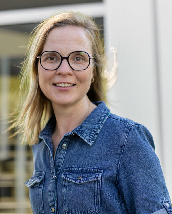 Lächelnde blonde Frau mit Brille. Sie trägt ein blaues Oberteil aus Denim.