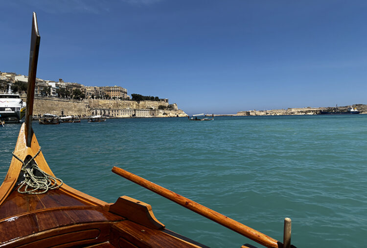 Maltesisches Boot, ähnlich einer venezianischen Gondel, auf grünblauem Meer. Im Hintergrund ein Ausschnitt von Valletta.