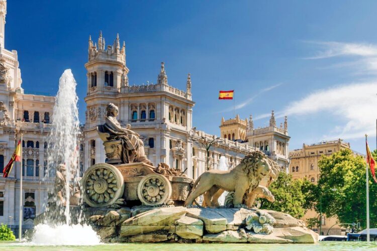 Ein Gebäude mit vielen kleinen Türmchen aus hellem Stein. Auf dem Dach weht die spanische Nationalflagge. Im Vordergrund ein großer Brunnen mit einer Kutsche und zwei Löwen aus Stein in der Mitte. Der Himmel ist strahlend blau.