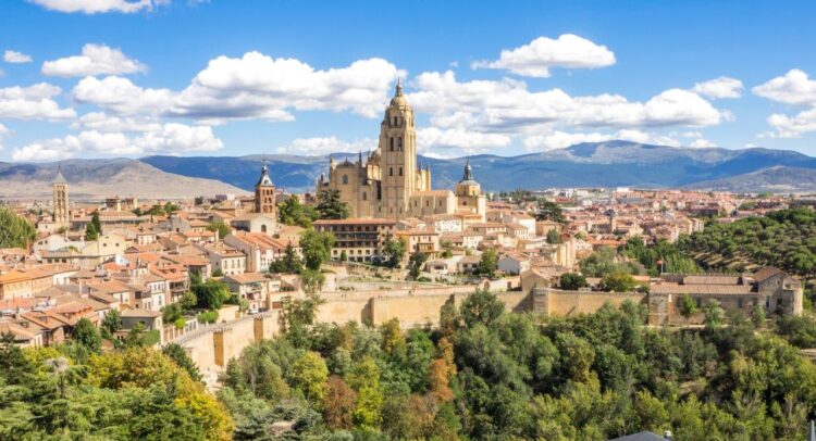 Kirche in Segovia in Spanien. Drumherum sind helle Gebäude und im Hintergrund eine hügelige bis bergige Landschaft.