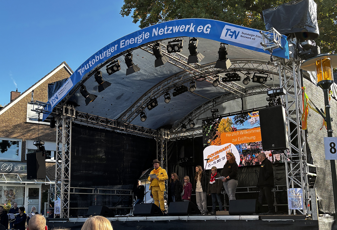 TEN-Bühne auf dem Ockermarkt. Ein ganz in gelb gekleideter Mann steht auf der Bühne und spricht ins Mikrofon. Im Hintergrund ist strahlend blauer Himmel zu sehen.