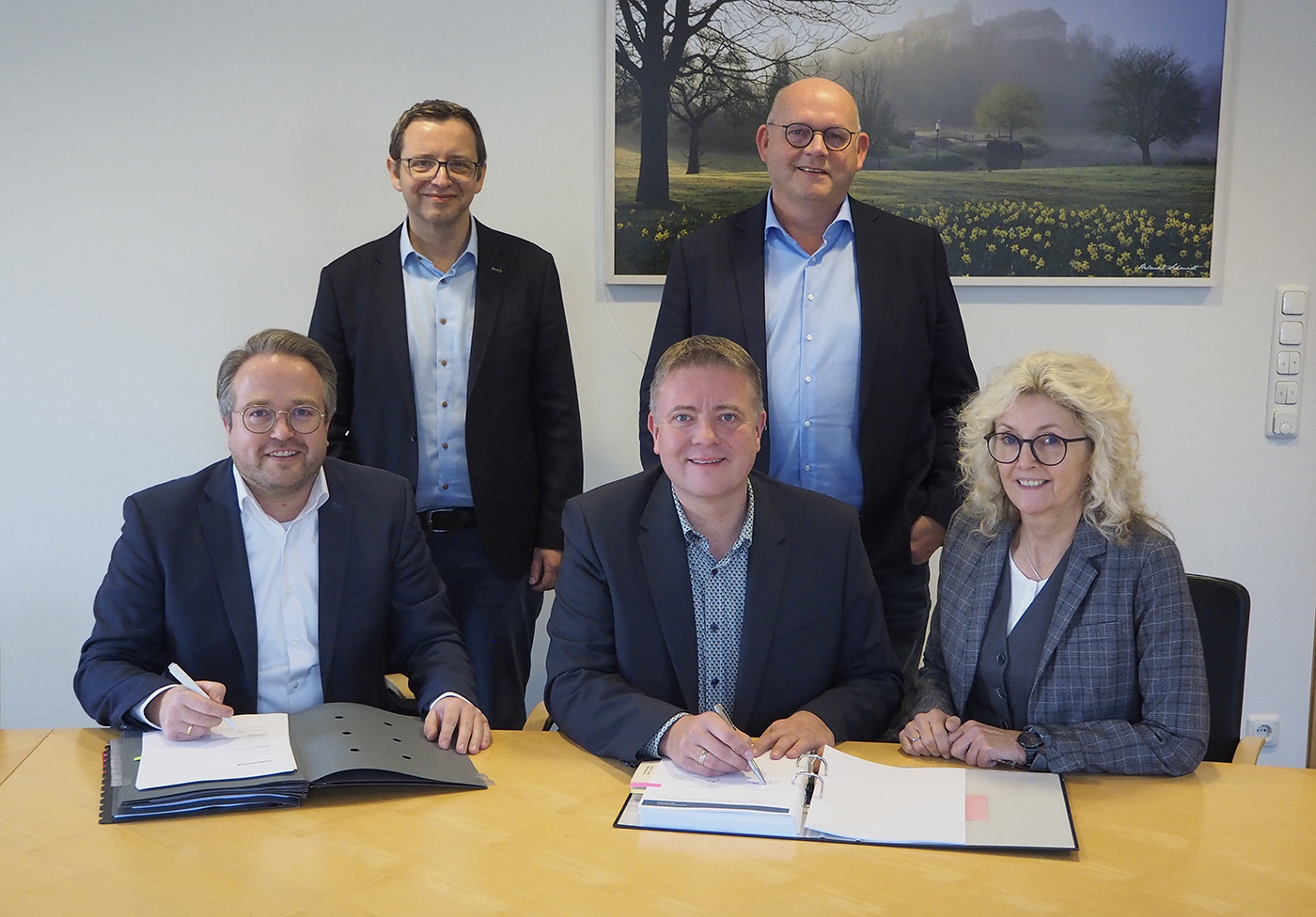 Zwei Männer und eine Frau sitzen nebeneinander an einem Tisch und unterzeichnen verschiedene Dokumente. Hinter den den drei Personen stehen zwei Männer und lächeln.